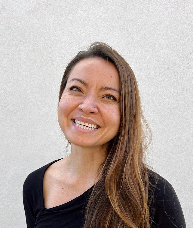 Smiling woman with long brown hair wearing a black top, standing against a light textured wall. She tilts her head slightly to the right and shows teeth.