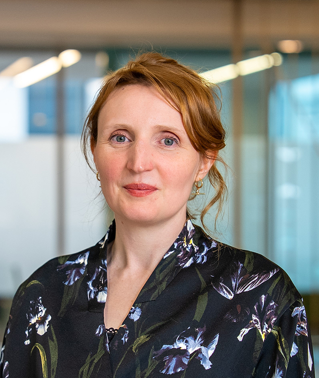 A portrait of a woman with light red hair wearing a dark floral blouse, standing indoors with glass walls in the background, soft smile, gold hoop earrings.