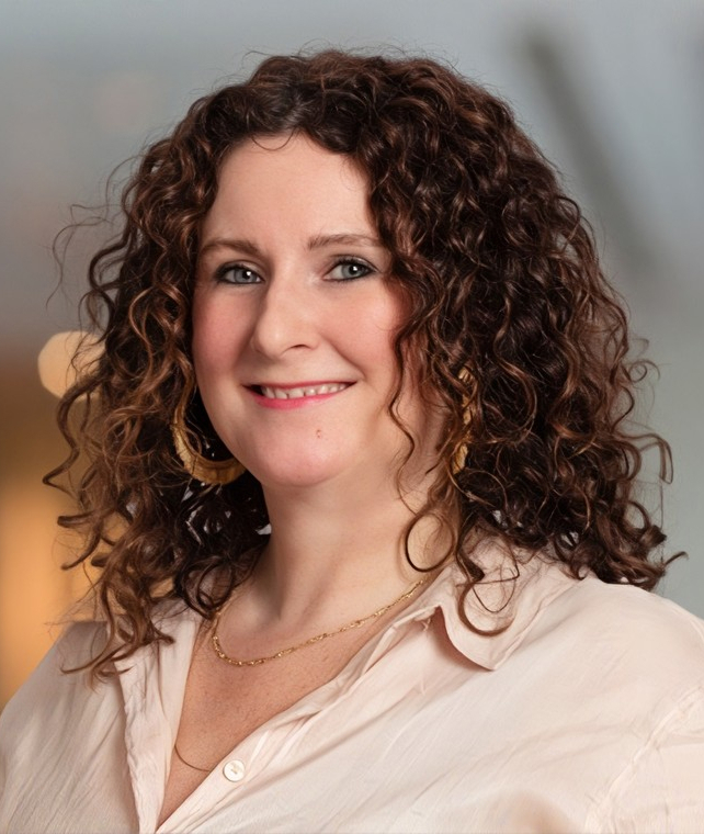 Portrait of a smiling woman with curly brown hair, light makeup, wearing a pale pink blouse and gold jewelry, softly lit indoor background.