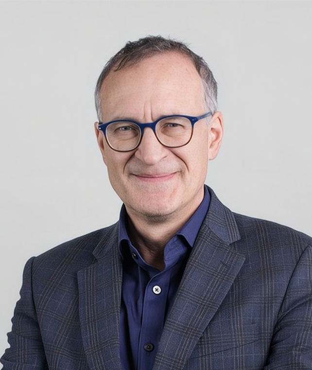 Portrait of a middle-aged man with short gray hair and blue framed glasses, wearing a dark blue checkered blazer over a navy shirt, smiling softly.