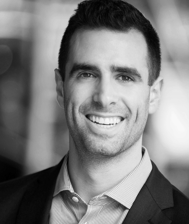 Smiling man in a suit jacket and checkered shirt, facing the camera with a friendly expression, short dark hair, and a blurred urban background.
