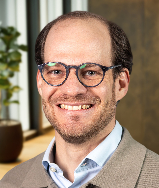 Smiling person with round blue glasses and light stubble, wearing a light blue collared shirt and beige jacket, indoor background with greenery.