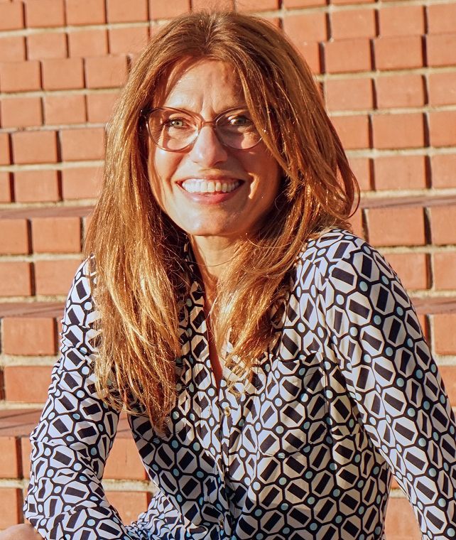 A smiling woman with long light brown hair and glasses sits outdoors in front of a brick staircase, wearing a geometric patterned blouse.