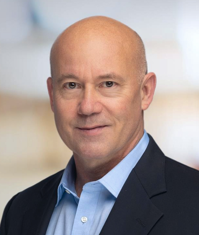 Professional headshot of a bald, middle-aged man in a blue dress shirt and dark blazer, neutral expression, soft indoor background.