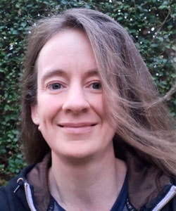 Portrait of a smiling woman with light brown hair outdoors, gentle breeze blowing strands, wearing a dark jacket, with green foliage in the background.