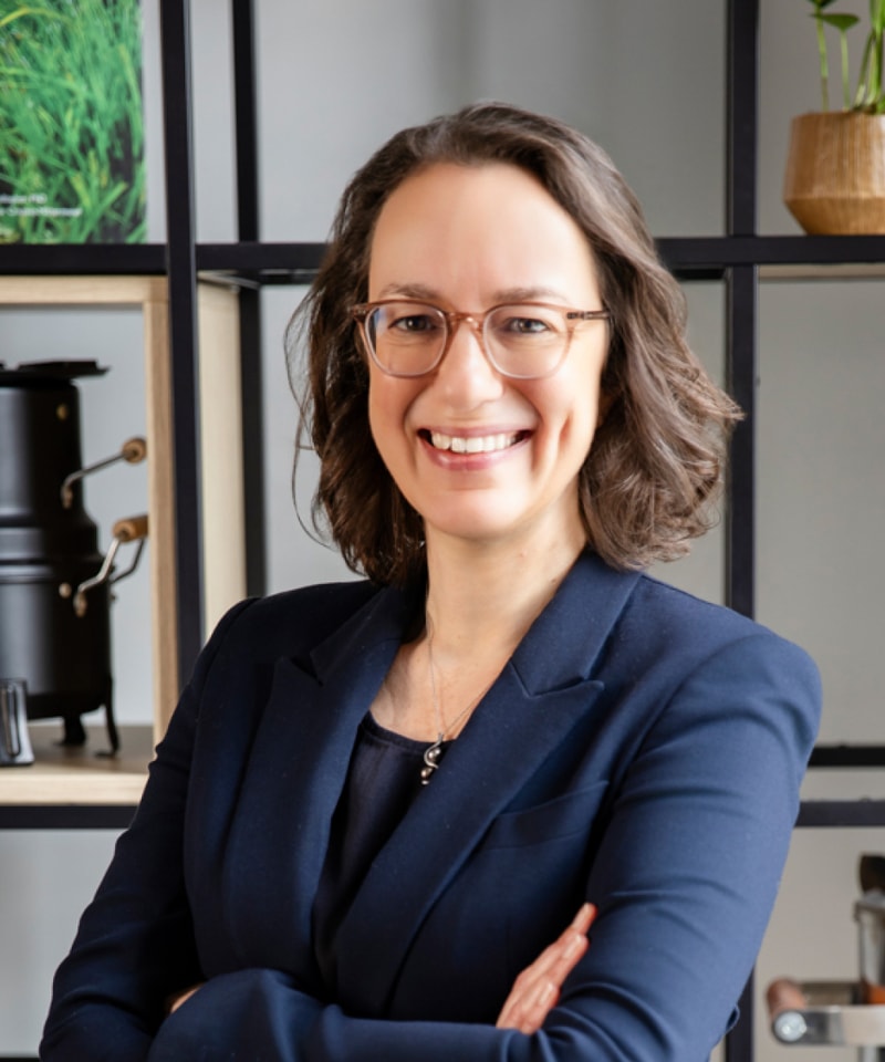 Smiling woman with short brown hair and glasses wearing a navy blazer, arms crossed, standing in a modern office with shelves and plants behind her.