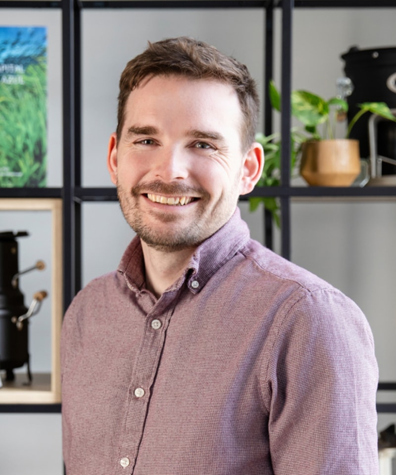 Smiling man with short brown hair and light beard wearing a maroon button-up shirt, standing in front of a shelf with plants and decor.