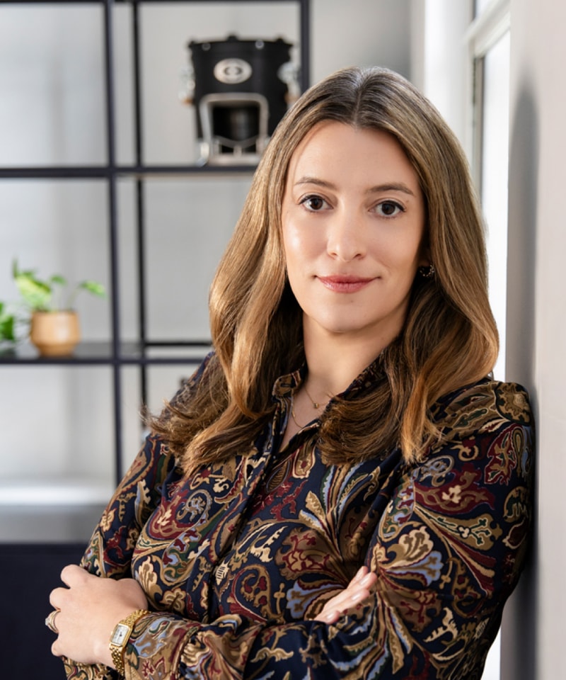 Professional woman with light brown hair stands with arms crossed in a modern office, wearing a patterned blouse and a gold watch, shelves and a coffee machine in the background.