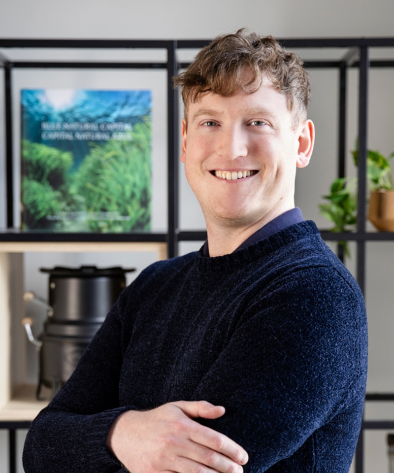 Smiling man with short light brown hair wearing a dark blue sweater stands with arms crossed in front of a shelving unit and framed greenery poster in a bright indoor setting.