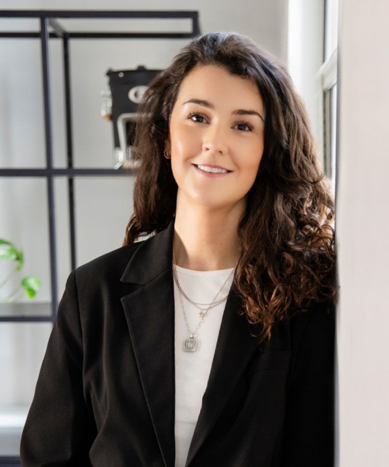 Smiling woman with wavy brown hair leans against a light wall in a modern office, wearing a black blazer over a white top and layered necklaces.