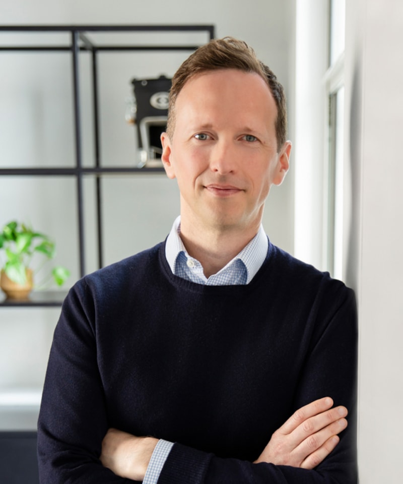 Portrait of a smiling man with short light brown hair, wearing a navy sweater over a light blue checkered shirt, standing with arms crossed indoors near a window.