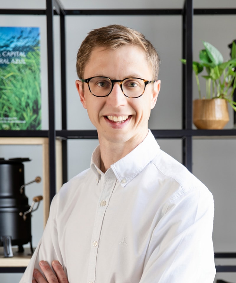 A smiling person with short light brown hair and round glasses wearing a white button-up shirt, standing in a modern office with shelves, plants, and framed posters in the background.
