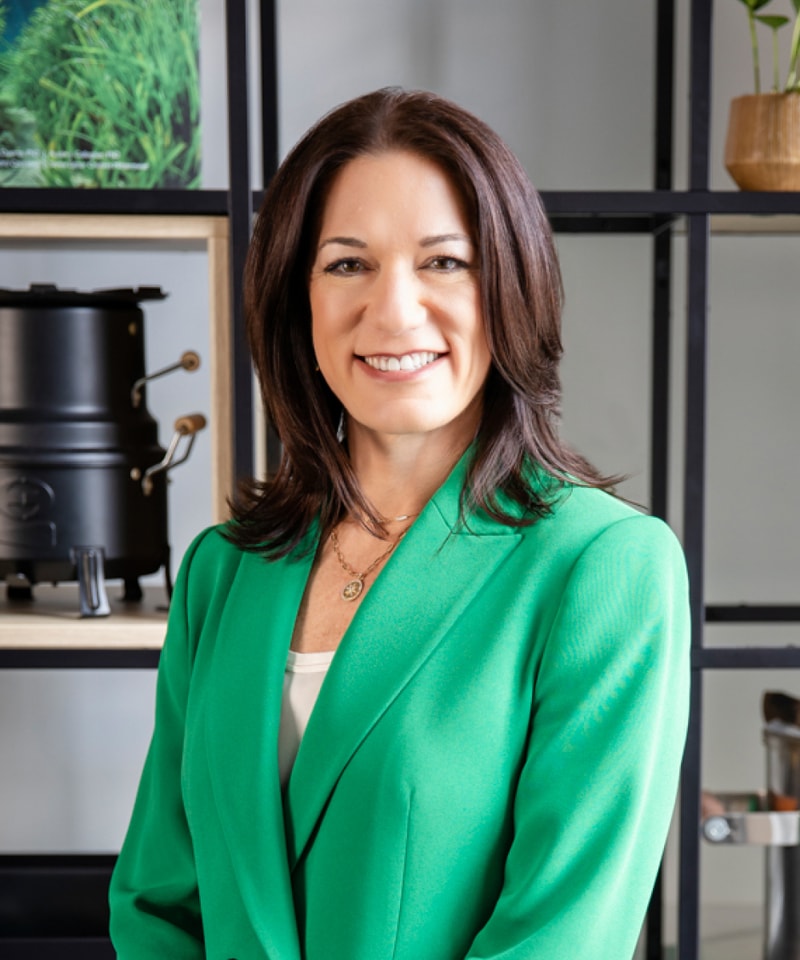 Smiling woman with shoulder-length dark brown hair wearing a green blazer and white top, standing in front of a shelving unit with decor and equipment.