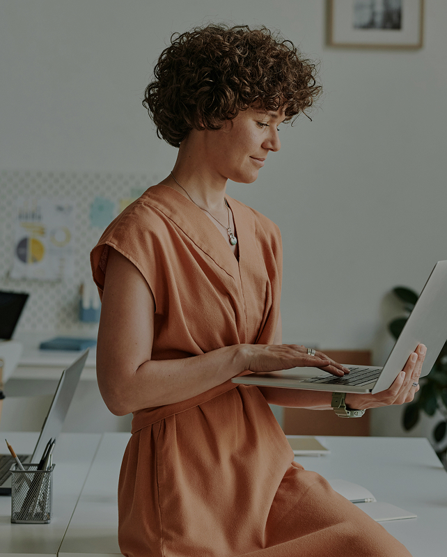 woman with short curly hair wearing a peach dress, standing at a desk and using a laptop in a bright, modern office space.