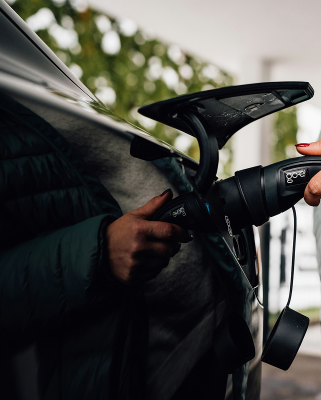 Person holding a portable car vacuum or handheld device attached to a car door, with a green jacket sleeve visible and a blurred outdoor background.