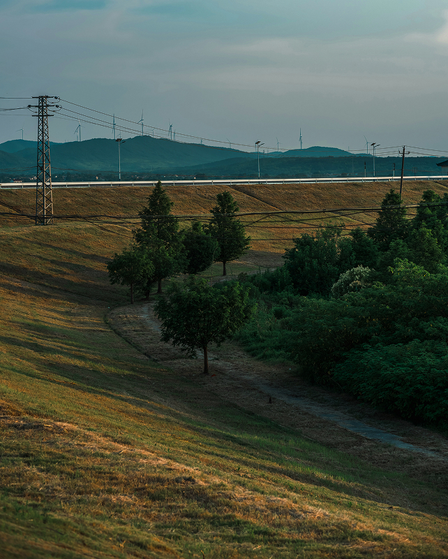 Hilly landscape with a dirt path and scattered trees in the foreground, a row of power lines and a highway in the midground, and wind turbines on distant hills under a cloudy sky.