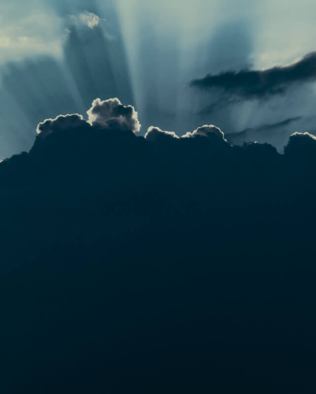 Dark clouds cover the lower portion with a bright sky above, illuminated by sharp sun rays breaking through the clouds from behind.