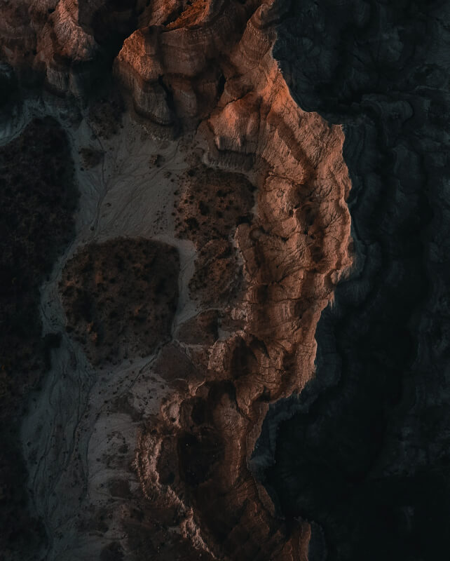 Aerial view of rugged coastline with reddish rock formations along a jagged cliff edge, casting deep shadows over textured terrain and dark sea below.