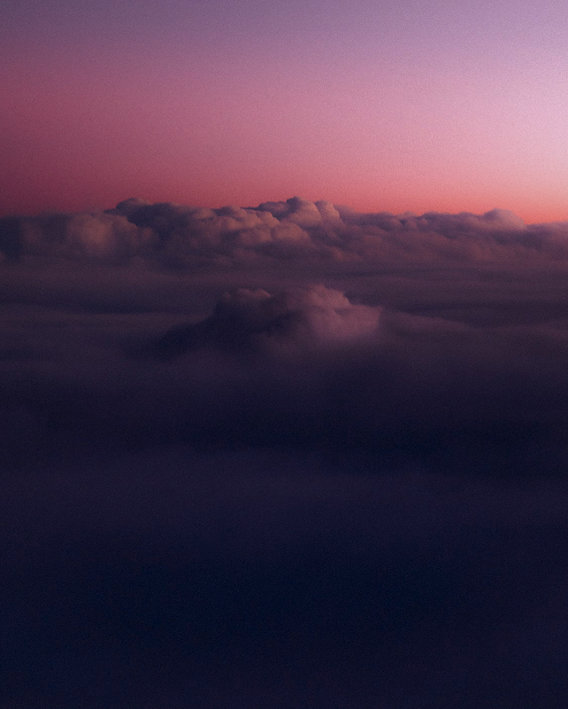 Vast, soft clouds float above a dark sea of lower clouds, with a pink and purple sunset sky casting gentle hues across the horizon.