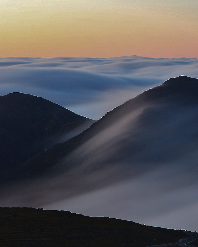 Soft pastel sunrise over misty mountain peaks, with dark ridges cutting through a swirling sea of fog and smooth, flowing clouds blanketing the valley.