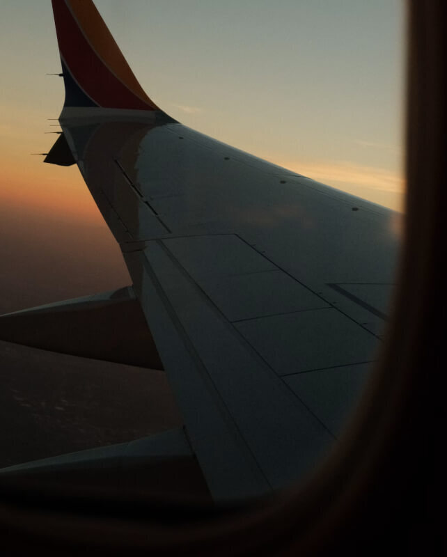 Wingtip and fuselage of an aircraft seen from a window, silhouetted against a sunset sky with warm orange hues, darkened wing panels, and window frame edges.