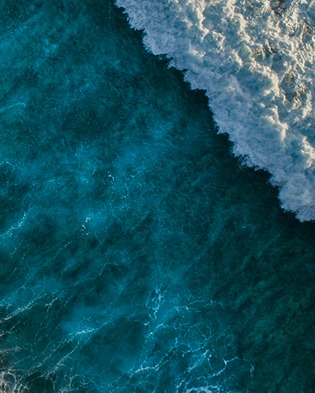 Aerial view of a turquoise ocean with white foamy waves crashing along the shore, showing textured water and light sea spray.