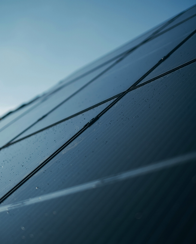 Close-up of sleek dark solar panels angled toward the sky, droplets of water on the glassy surface, with a blue gradient sky in the background.
