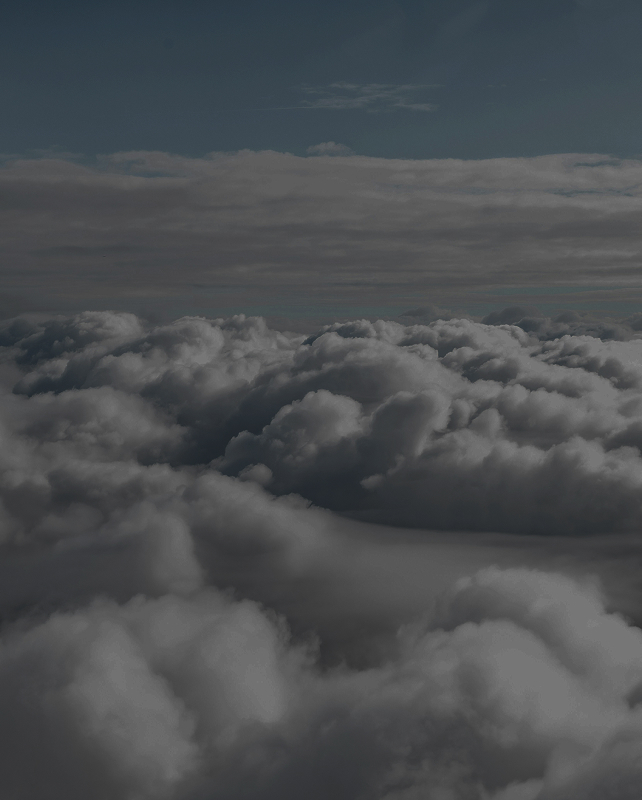 Clouds drifting above and below, with a distant blue sky and thin high clouds near the horizon, creating a moody, layered cloudscape viewed from above.