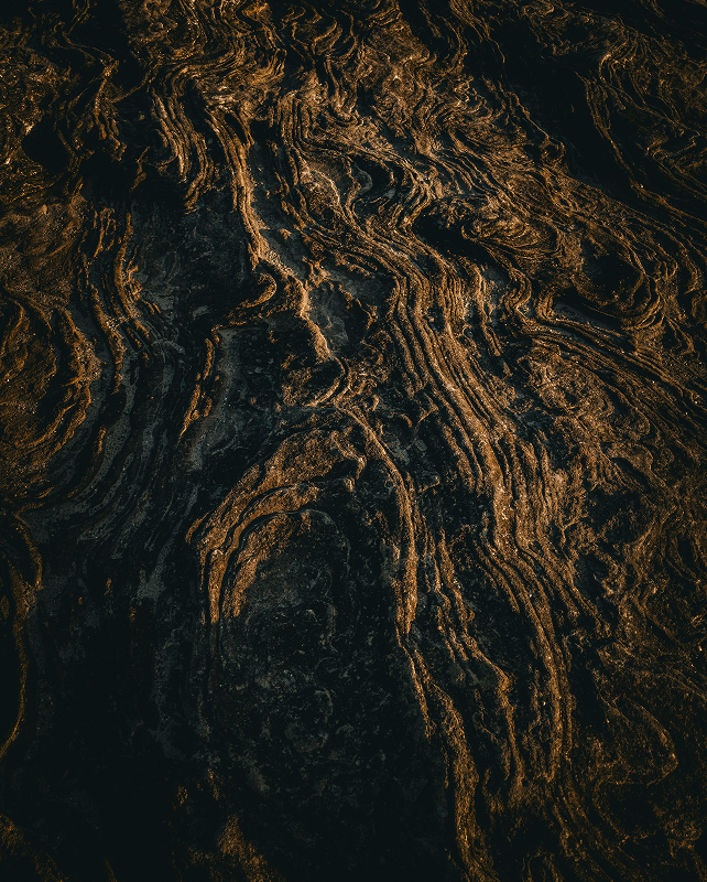 Dark, textured lava or rock surface with winding, golden-brown striations and ridges creating a rugged, abstract pattern.